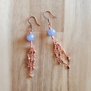 Puede incluir: Un par de pendientes de cobre con una cuenta de piedra preciosa azul claro y una cadena colgante de cuentas de cobre.