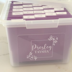 May include: A clear plastic storage box with a purple lid and a purple interior. The box is labeled with the names "Presley Tayhsa" and has dividers labeled with different age groups and grades, including "Baby", "1-2 Years", "2-3 Years", "3-4 Years", "Preschool", "Pre-K", "Kindergarten", "1st Grade", "2nd Grade", "3rd Grade", "4th Grade", and "5th Grade".