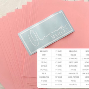 May include: A stack of pink cardstock paper with a white label on top. The label has the name "Olivia Marley" written in a cursive font. The cardstock is divided into columns with headings for different categories, such as Pregnancy, Baby, 1-2 Years, 2-3 Years, 3-4 Years, Preschool, Pre-K, Kindergarten, 1st Grade, 2nd Grade, 3rd Grade, 4th Grade, 5th Grade, 6th Grade, 7th Grade, 8th Grade, 9th Grade, 10th Grade, 11th Grade, 12th Grade, Graduation, College, School Info, Report Cards, Certificates, Awards, Achievements, School Pictures, Photos, Documents, Sports, Dance, Extracurricular, Artwork, Keepsakes, Birthday Cards, Health Records, Medical, Financial, and Misc.