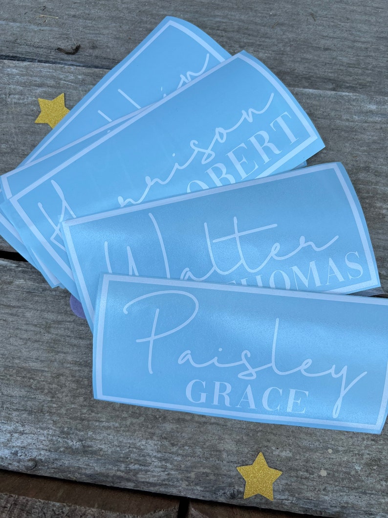Pu&ograve; includere: Un set di adesivi per nomi personalizzati di colore azzurro chiaro con lettere bianche. I nomi sono "William", "Harrison", "Robert", "Walter", "Thomas", "Paisley" e "Grace".