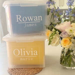 May include: Two clear plastic storage boxes with yellow and blue lids. The boxes are labeled with the names "Rowan James" and "Olivia Marie".