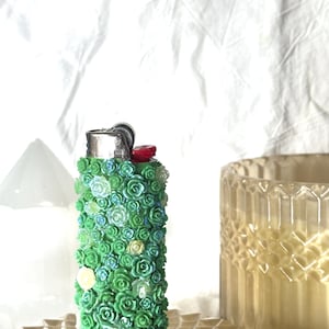 May include: A decorative lighter covered in small, green, blue, and white floral embellishments. The lighter has a silver metal top and a red ignition button. The lighter is on a decorative, light-colored surface.