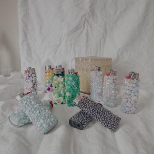 May include: A collection of decorative lighters in various colors and designs. Some lighters are embellished with beads, pearls, and other decorative elements. One lighter has the text "HUG ME". The lighters are arranged on a white surface.