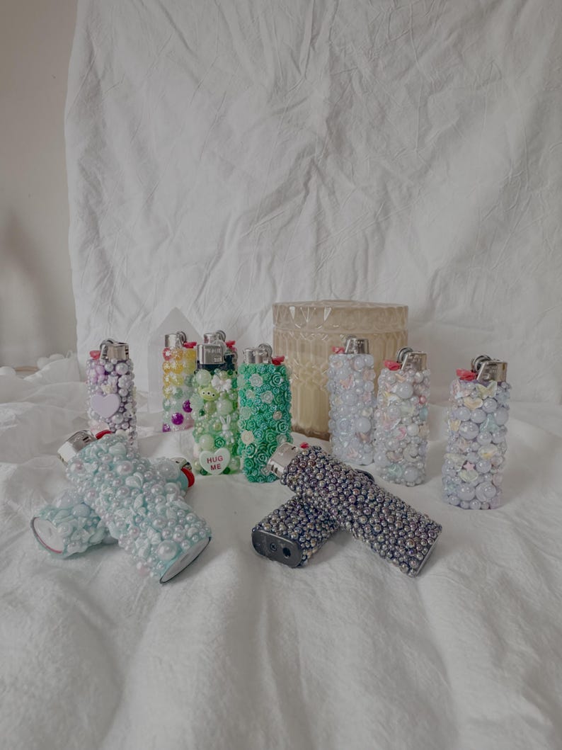 May include: A collection of decorative lighters in various colors and designs. Some lighters are embellished with beads, pearls, and other decorative elements. One lighter has the text "HUG ME". The lighters are arranged on a white surface.