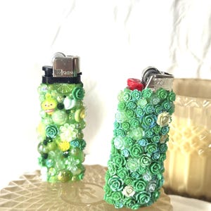 May include: Two decorative lighters. One is covered in green and yellow beads and charms. The other is covered in small green roses. Both lighters have silver metal tops and are on a clear, textured surface.