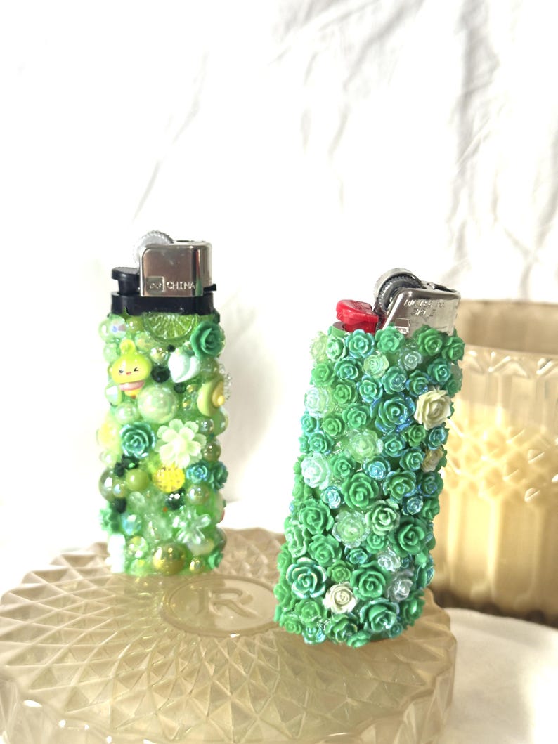 May include: Two decorative lighters. One is covered in green and yellow beads and charms. The other is covered in small green roses. Both lighters have silver metal tops and are on a clear, textured surface.