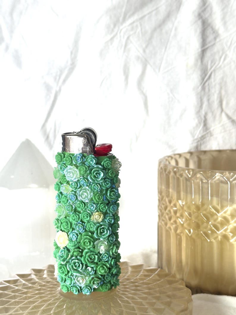 May include: A decorative lighter covered in small, green, blue, and white floral embellishments. The lighter has a silver metal top and a red ignition button. The lighter is on a decorative, light-colored surface.