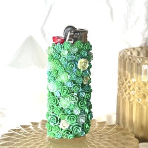 May include: A decorative lighter covered in small, green, blue, and white roses. The lighter has a silver metal top and a red button. The roses are arranged closely together, creating a textured surface. The lighter is standing on a clear, patterned surface.