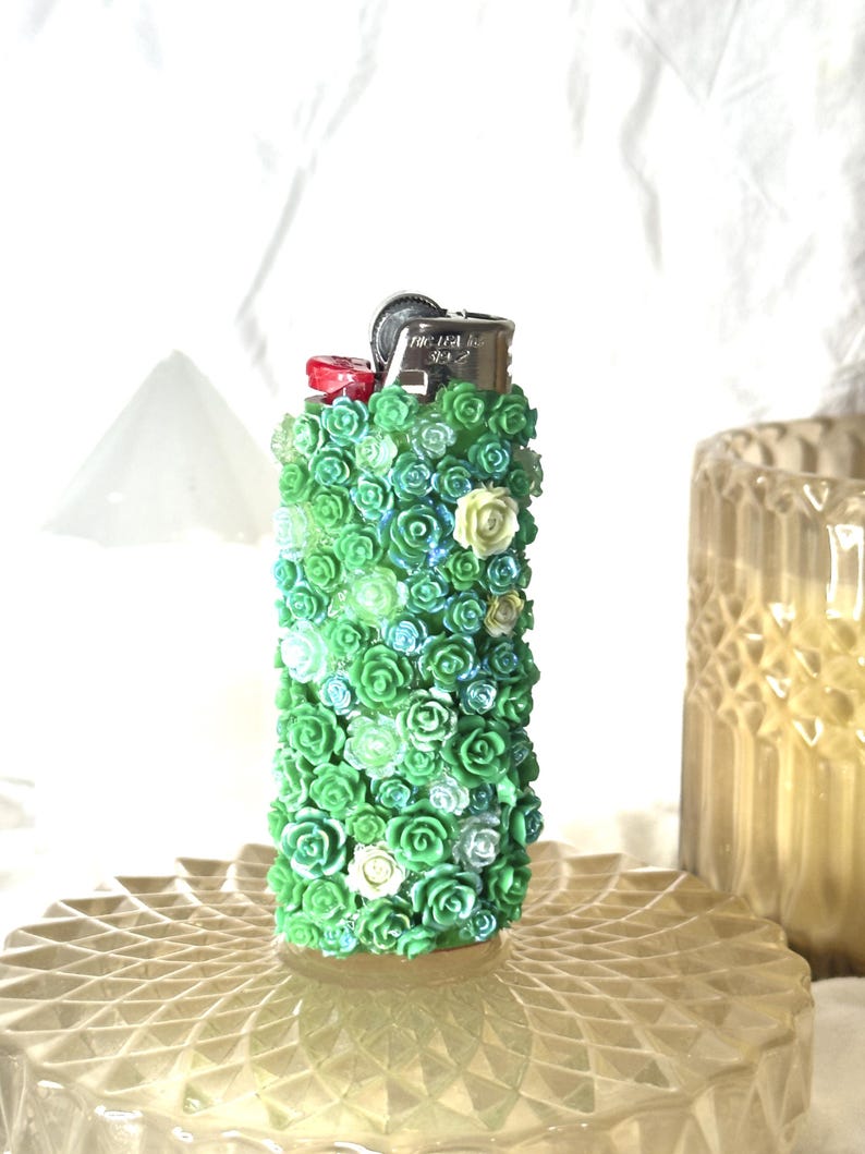 May include: A decorative lighter covered in small, green, blue, and white roses. The lighter has a silver metal top and a red button. The roses are arranged closely together, creating a textured surface. The lighter is standing on a clear, patterned surface.