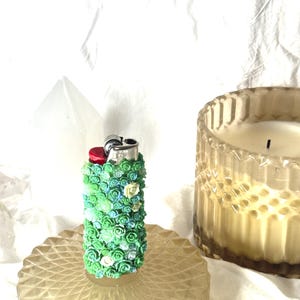 May include: A decorative lighter covered in small, colorful flowers in shades of green, blue, and white. The lighter sits on a round, textured, amber-colored glass dish. A candle in a glass jar is in the background.