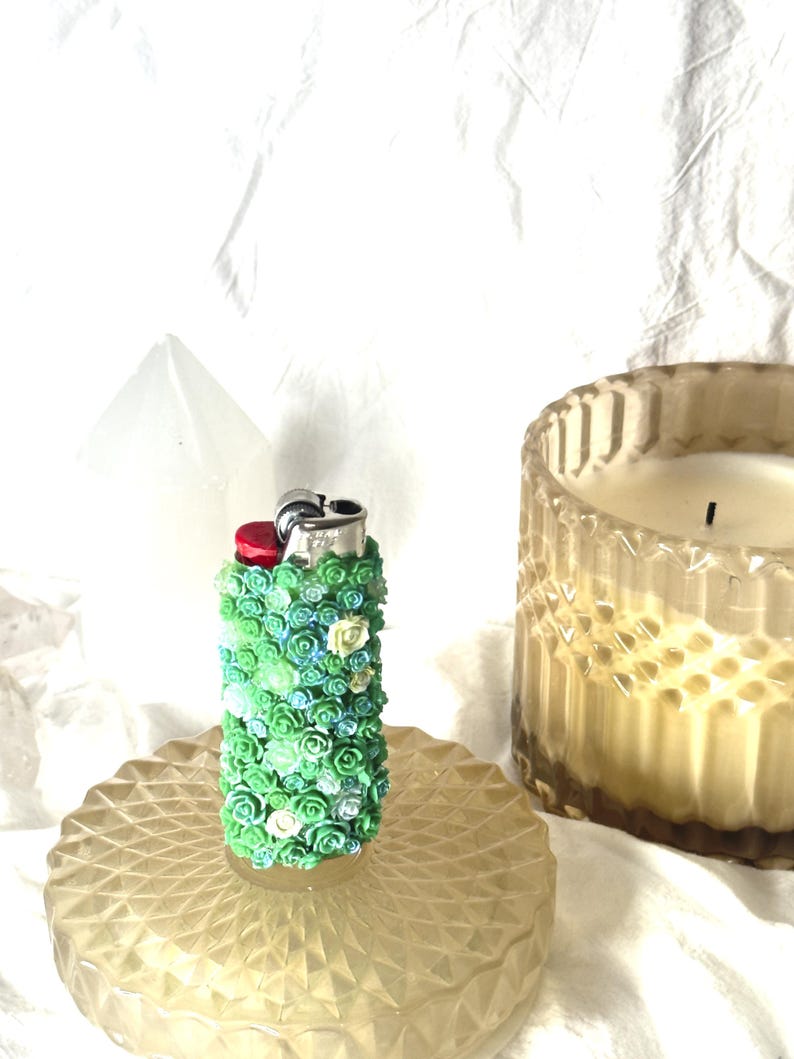 May include: A decorative lighter covered in small, colorful flowers in shades of green, blue, and white. The lighter sits on a round, textured, amber-colored glass dish. A candle in a glass jar is in the background.