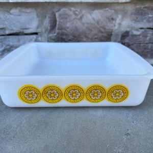 May include: A white, square Pyrex baking dish with rounded corners. The dish is decorated with five yellow and brown floral medallions along the front. The dish is sitting on a gray surface.