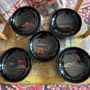 May include: Five black lacquerware dishes with a Japanese landscape design. Each dish features a red sun, a torii gate, and stylized birds and mountains in gold and white. The dishes are round and have a glossy finish.