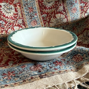 May include: Two white ceramic bowls with a scalloped edge and a green trim are stacked on top of each other. The bowls are set against a colorful patterned fabric with red, blue, and cream tones. The bowls are likely for serving food.