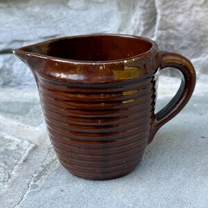 May include: A brown ceramic pitcher with a handle and a spout. The pitcher has a ribbed texture around the body. The interior is a darker brown. The pitcher is sitting on a stone surface.
