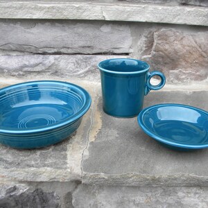 May include: A set of three teal-colored ceramic dishes. The set includes a large oval bowl, a small oval dish, and a mug with a handle. The dishes have a glossy finish and are arranged on a stone surface.