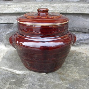May include: A brown ceramic casserole dish with a lid. The dish has a glossy finish and decorative wave and floral patterns. The lid has a knob and a gold-colored rim. The dish has two handles on the sides.