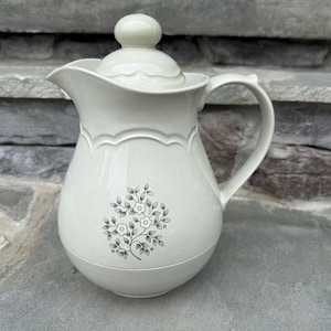 May include: Cream-colored ceramic pitcher with a lid and handle. The pitcher features a floral design and scalloped edges. The lid has a small, round knob. The pitcher is sitting on a stone surface.