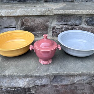 May include: Three ceramic serving pieces in shades of yellow, dusty rose, and gray. The set includes two large, shallow bowls and a lidded sugar bowl with handles. The bowls have a ridged design, and the sugar bowl has a pedestal base.