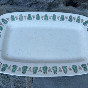 May include: Rectangular serving platter with a speckled off-white surface. The rim is decorated with a repeating pattern of green and brown stylized triangles. A vintage-style piece for serving food.