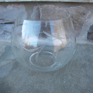 May include: A clear glass bowl with etched floral designs. The bowl has a rounded shape and sits on a stone surface. The etched flowers and leaves add a decorative touch to the glassware.