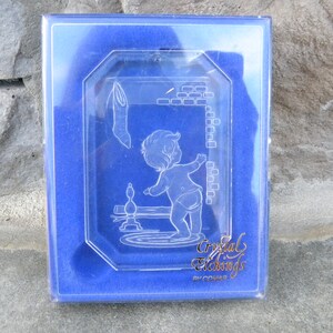 May include: A clear, etched glass ornament featuring a baby reaching towards a hanging stocking. The ornament is in a blue box with the words "Crystal Etchings" in gold lettering.