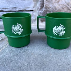 May include: Two green plastic mugs with a white bird and leaf design. Each mug has a handle and a base that fits into the bottom of the mug. The mugs are sitting on a gray surface.