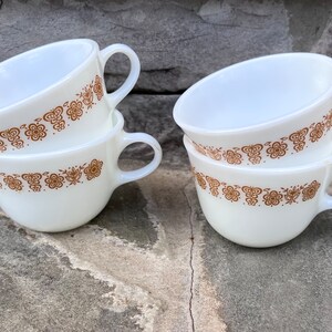 May include: Four white ceramic cups with a brown floral pattern around the rim. Each cup has a handle and is stacked in pairs. The cups are set against a textured gray stone background.
