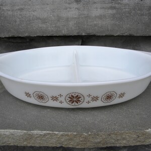 May include: White oval divided casserole dish with brown floral and geometric patterns. The dish has two handles and a divider in the center. A vintage kitchenware item.