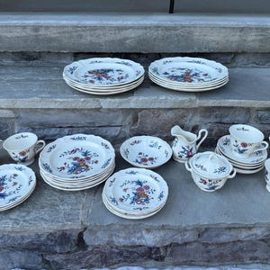 May include: A collection of vintage porcelain dishware with a floral design in red, blue, and yellow. The set includes plates, bowls, cups, saucers, a creamer, and a covered serving dish. The edges of the plates and bowls have a scalloped design.