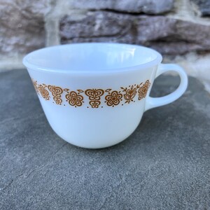 May include: White teacup with a brown floral pattern around the rim. The cup has a handle and is made of ceramic. The floral design includes flowers and butterfly shapes. The cup is sitting on a gray surface.