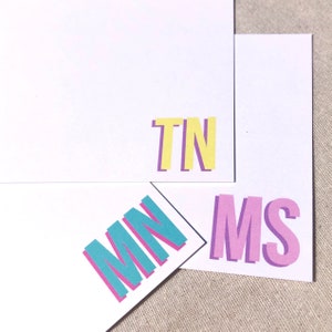 May include: Three cards with different colored letters on each. The first card has the letters "TN" in yellow with purple outlines. The second card has the letters "MN" in turquoise with pink outlines. The third card has the letters "MS" in pink with purple outlines.