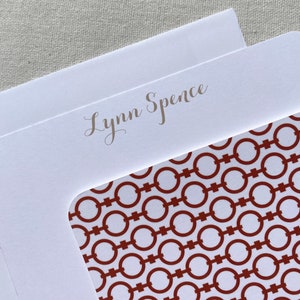 May include: White stationery card with a red and white geometric pattern on the bottom half. The top half has the name "Lynn Spence" written in silver script.