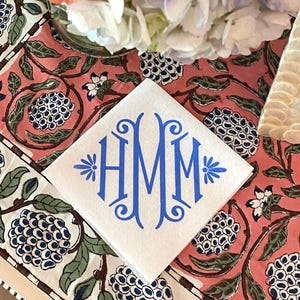 May include: A white linen napkin with a blue monogram of the letters "HMM" in a decorative script font. The napkin is on a pink and white floral tablecloth.