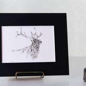 May include: A framed art print featuring a detailed black and white illustration of a stag with large antlers. The artwork is displayed in a black mat and frame, resting on a brass easel. The word "SAMPLE" is printed at the bottom.