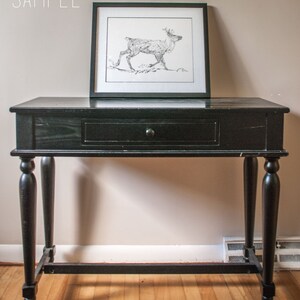 May include: Black console table with a single drawer and turned legs. A framed black and white drawing of a reindeer rests on the table. The word "SAMPLE" is in the upper left corner.