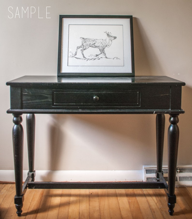 May include: Black console table with a single drawer and turned legs. A framed black and white drawing of a reindeer rests on the table. The word "SAMPLE" is in the upper left corner.