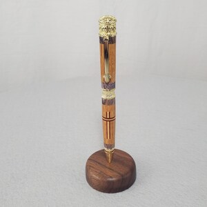 May include: A handcrafted wooden pen featuring alternating light and dark wood segments with gold-toned metal accents, including a decorative top finial, a central band, and a clip, standing upright in a circular dark wood base.