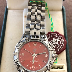 May include: A silver wristwatch with a red face and black hands. The watch has a silver band and a silver bezel with black numbers. The watch face also has the words "Quartz Water Resistant" and "Japan Movt". The watch is in a box with a red and white label that says "Hollywood Polo Sport".