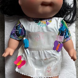 May include: A vintage Cabbage Patch Kid doll with black hair and blue eyes. The doll wears a white dress with butterfly accents and a blue and pink butterfly tag. The doll is lying on a white surface.