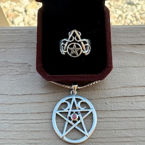 May include: A silver ring with a pentagram design inside a velvet jewelry box. Below, a silver pendant with a pentagram and crescent moon design, with a small red stone in the center, hangs on a chain.