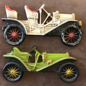 May include: Two vintage toy cars, one white with red accents and one green, both with black wheels and detailed features. The cars are made of metal and have a classic design.