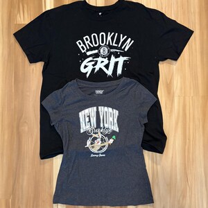 May include: Two t-shirts, one black and one gray, are displayed. The black shirt features the text "BROOKLYN GRIT" in white. The gray shirt has "NEW YORK Brooklyn" with a Looney Tunes character graphic.