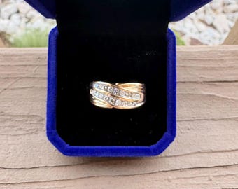 May include: A gold ring featuring two intertwined bands with a channel setting of small clear stones. The ring is displayed inside a dark blue velvet jewelry box with a black interior lining, highlighting the metallic luster and stone clarity.