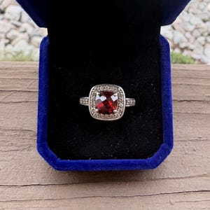May include: A silver-toned ring featuring a square-cut, deep red gemstone at the center, surrounded by a halo of small, clear, sparkling stones. The ring is presented inside a dark blue velvet-lined jewelry box with a black interior insert.