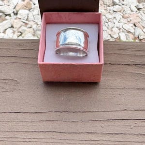 May include: A wide, polished silver band ring with a smooth, reflective surface. The ring is presented inside a small, square, light pink cardboard gift box with a white interior lining, designed to hold and display the jewelry piece securely.
