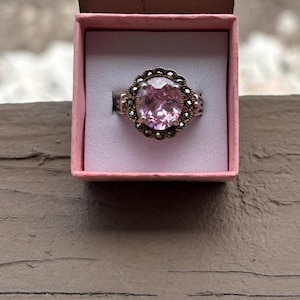 May include: A ring featuring a large, round, faceted light pink stone set in a dark metal frame surrounded by a halo of small, dark, circular stones, presented inside a small, square, light pink cardboard jewelry box with a white insert.