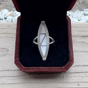 May include: Silver ring with a long, boat-shaped design. The ring features a white, inlaid stone with a geometric pattern. The ring is displayed in a maroon velvet ring box, showcasing the jewelry's design and craftsmanship.