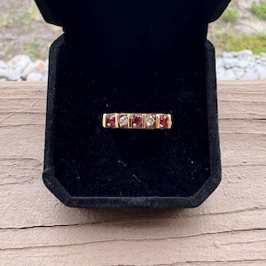 May include: A gold ring with alternating red and clear stones, presented in a black velvet box. The ring features a series of rectangular settings, each holding a small gemstone. The ring is displayed against a natural wood background.