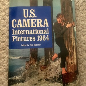May include: A vintage book cover featuring a woman in a black outfit standing on a wooden pier with water splashing around her. The book title is "U.S. CAMERA International Pictures 1964" and the text "Edited by Tom Maloney" is below the title.
